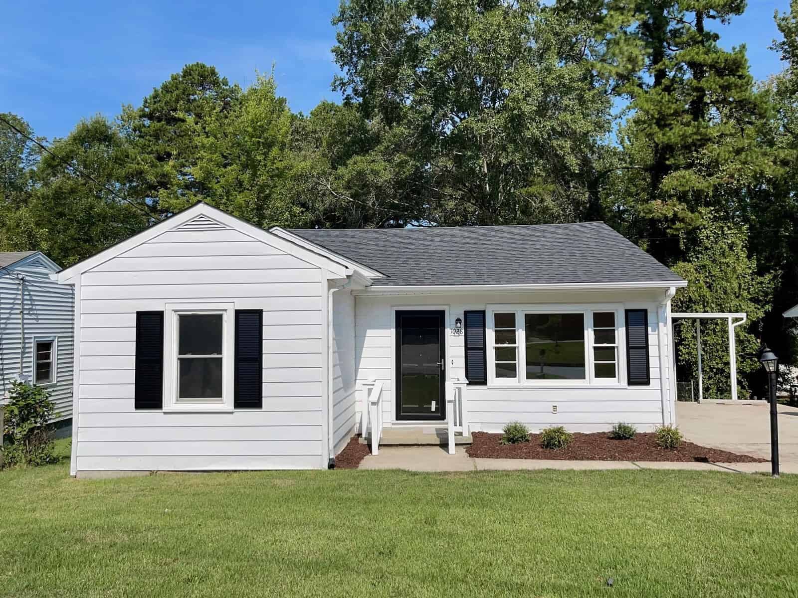 A small, white single-story house with a dark roof and green lawn is located at 1028 Roanoke Avenue, Henderson NC. The house features a small porch, two front windows, and