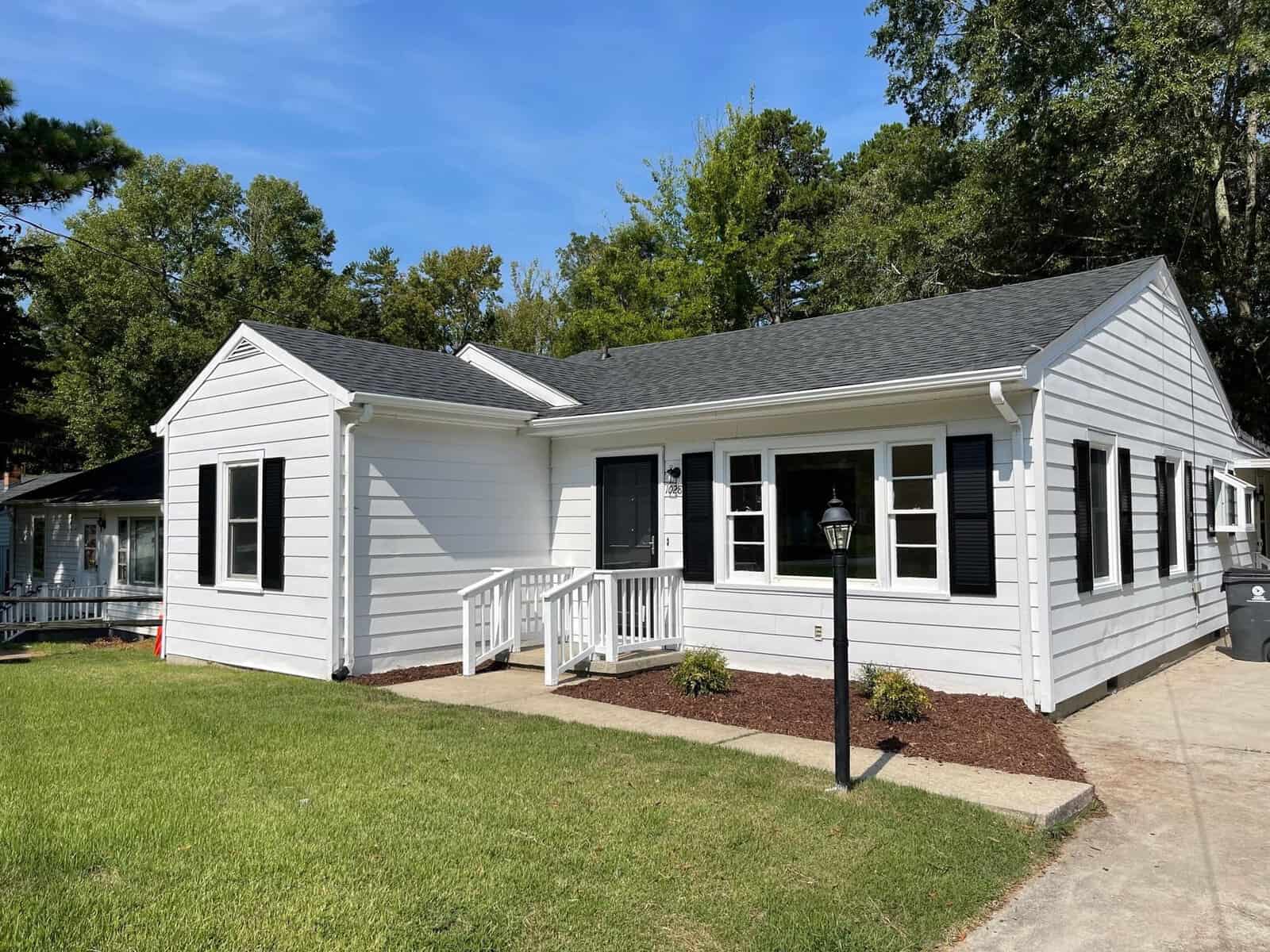 A single-story white house with black shutters, featuring a small porch and a front lawn. a lamppost and pathway lead to the entrance, set against a backdrop of trees and clear blue sky.