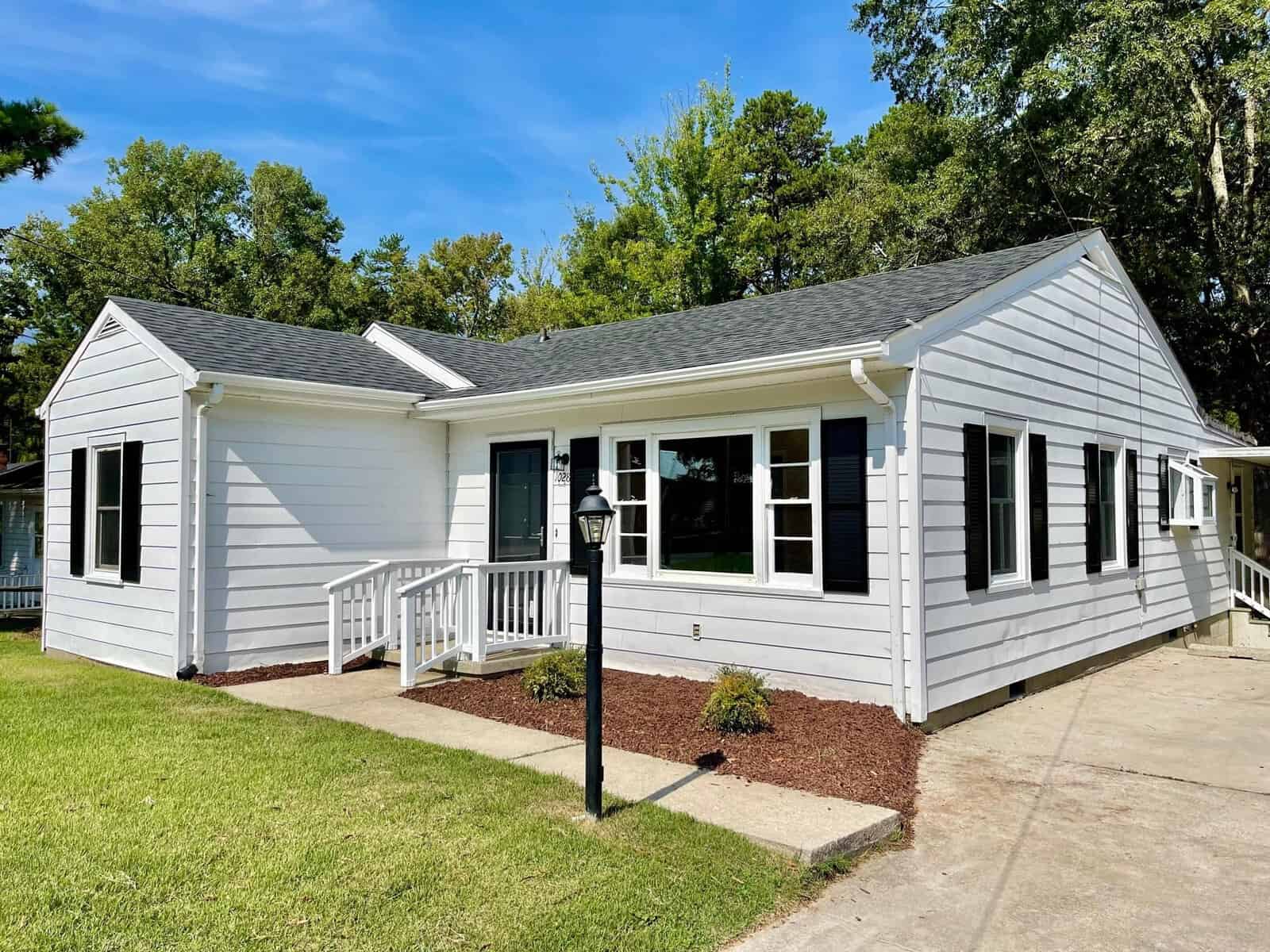 A freshly painted white single-story house with a gray shingle roof, surrounded by green trees and a neat yard, under a blue sky.