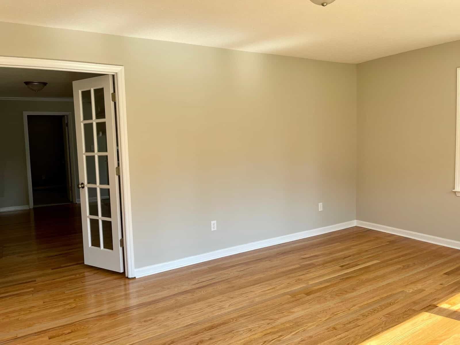 Empty room with hardwood floors and beige walls, featuring a white open french door leading to another room. bright natural light entering from the side.