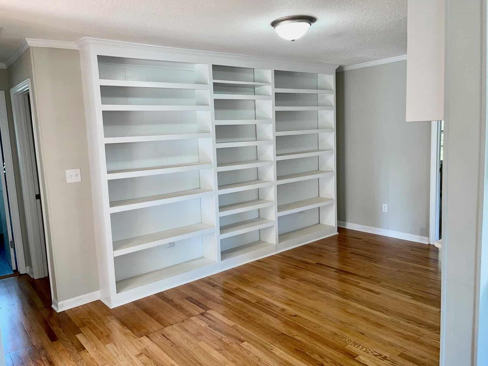 A bright room featuring an empty, large built-in white bookshelf along one wall with multiple shelves. the floor is covered with polished hardwood.