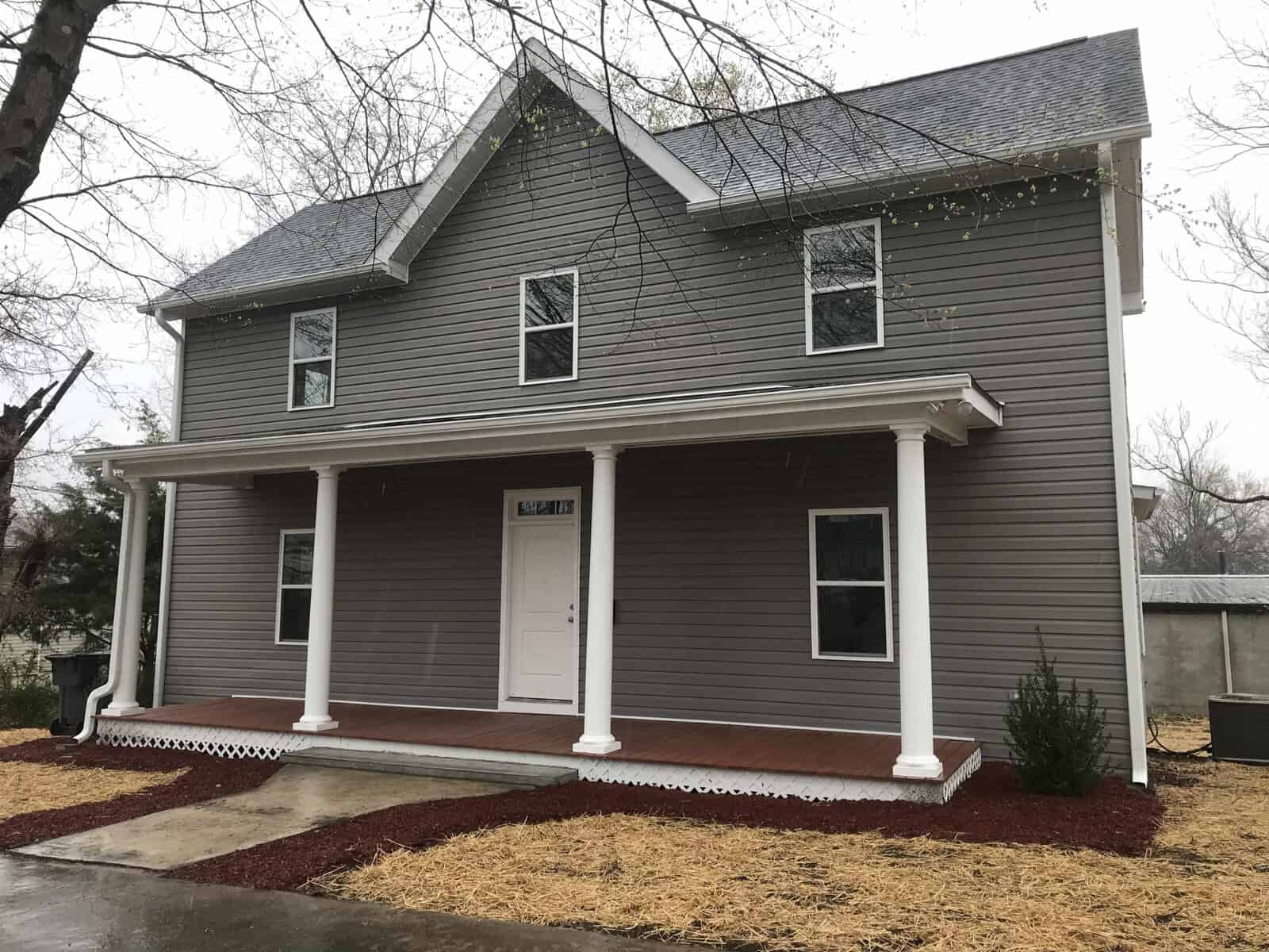 A two-story residential home with grey siding and white trims, featuring a covered front porch and a red pathway, under an overcast sky.