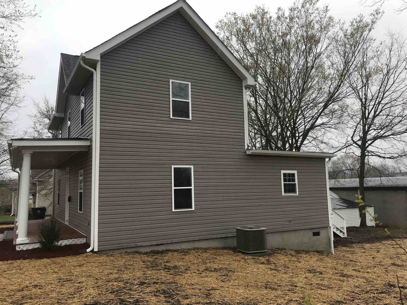 A two-story house with grey siding and white trim on an overcast day, featuring a covered porch and a barren yard with freshly laid straw mulch.