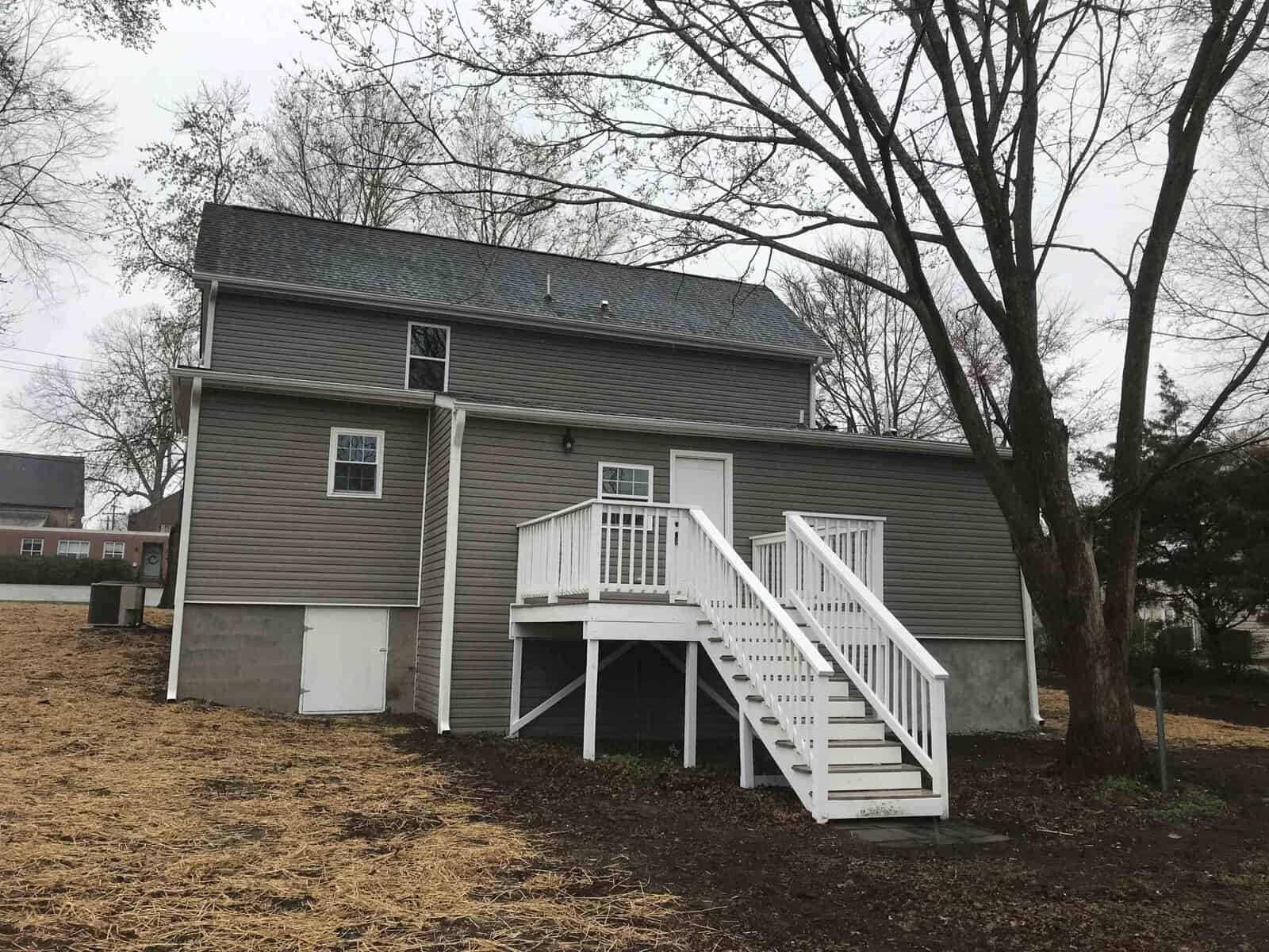 A two-story house with gray siding and white trim, featuring an upper-level deck with white stairs leading down to the ground. there are bare trees and a partially cloudy sky in the background.