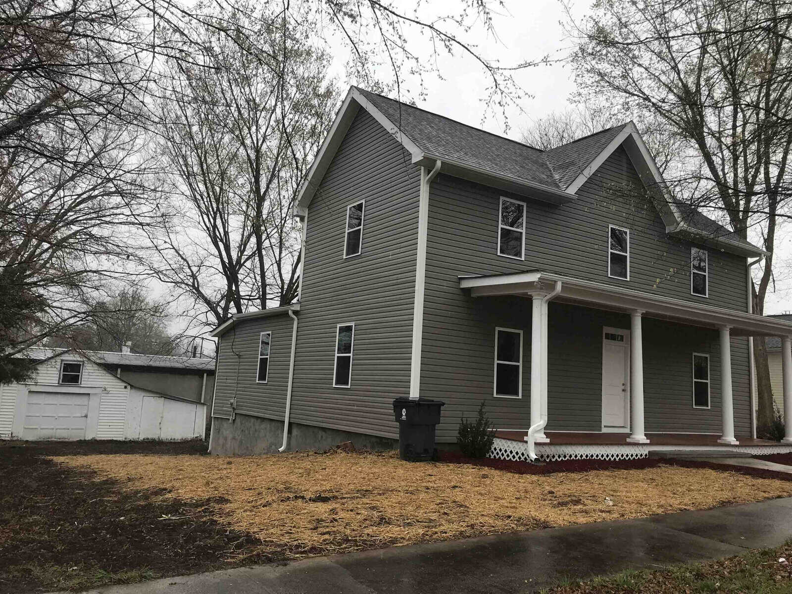 A two-story gray house with white trim, a covered porch, and a detached garage to the left of the property, all under an overcast sky.