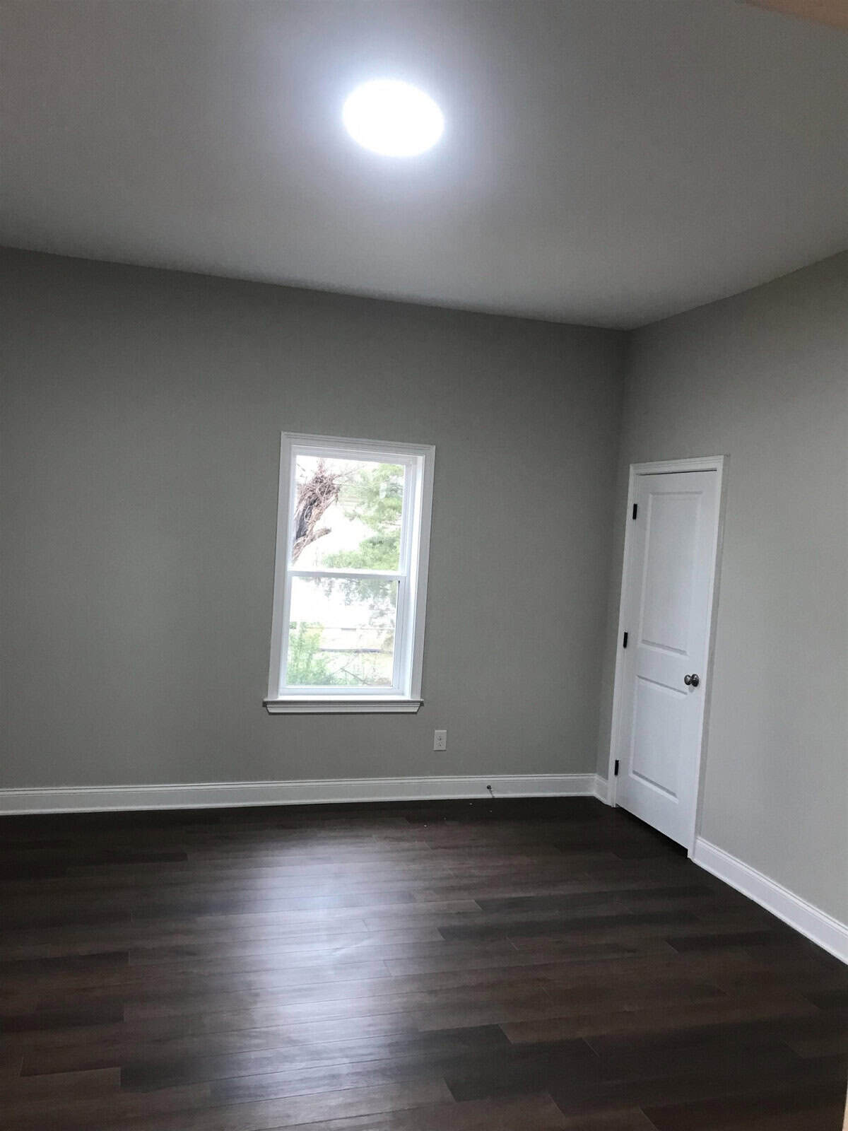 An empty room with gray walls, a single window letting in natural light, and dark hardwood flooring. there is a white door on the right side of the frame.