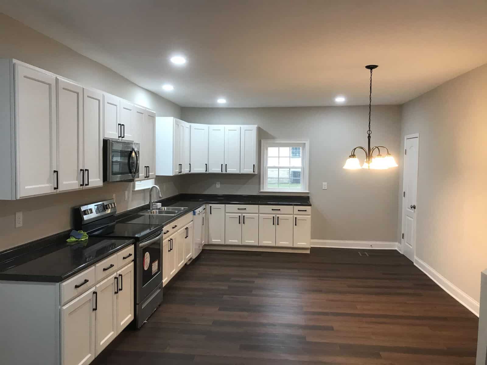 A modern kitchen with white cabinetry, dark countertops, stainless steel appliances, recessed lighting, and an adjoining dining area with a hanging light fixture.