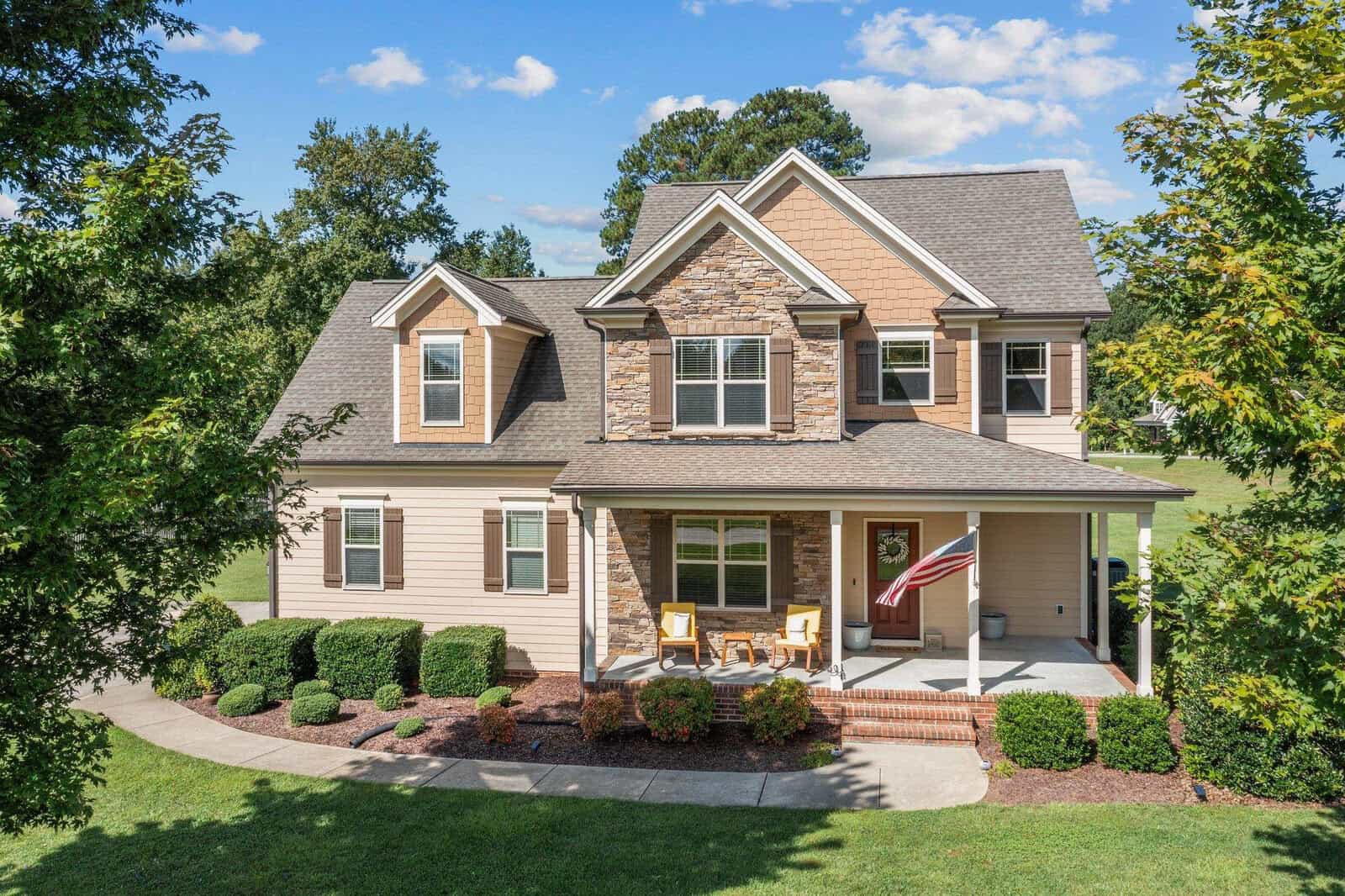 A two-story suburban house at 1721 Creekview Drive in Franklinton features a stone and siding facade, surrounded by a neatly maintained lawn and bushes. An American flag hangs by the front porch, where two wooden chairs are placed. The sky is clear and dotted with a few clouds.