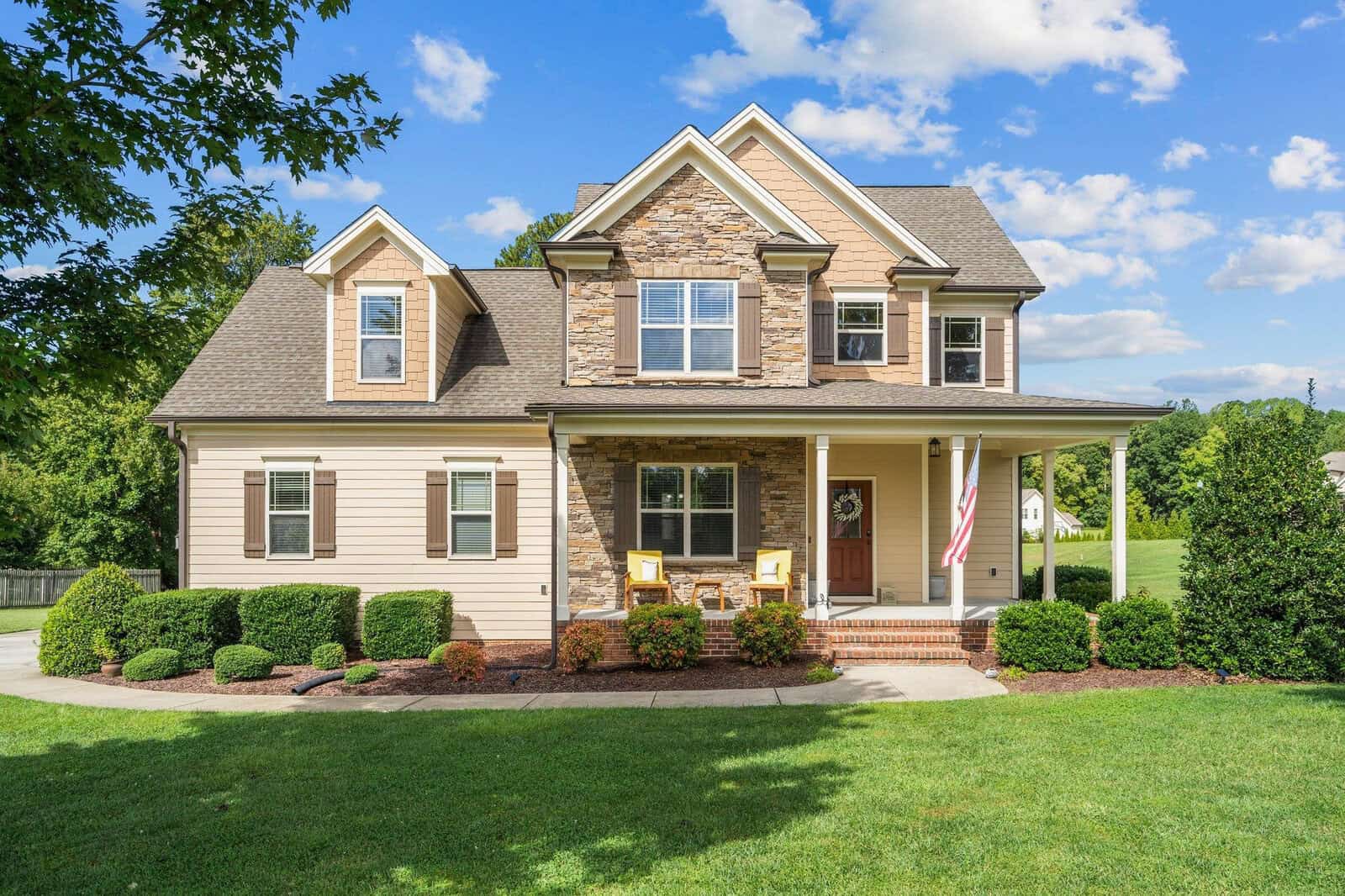 A two-story suburban house with stone and siding exterior, a gabled roof, and a welcoming front porch. Two chairs and an American flag decorate the porch. The yard is well-maintained with green grass and bushes, and the sky is partly cloudy.