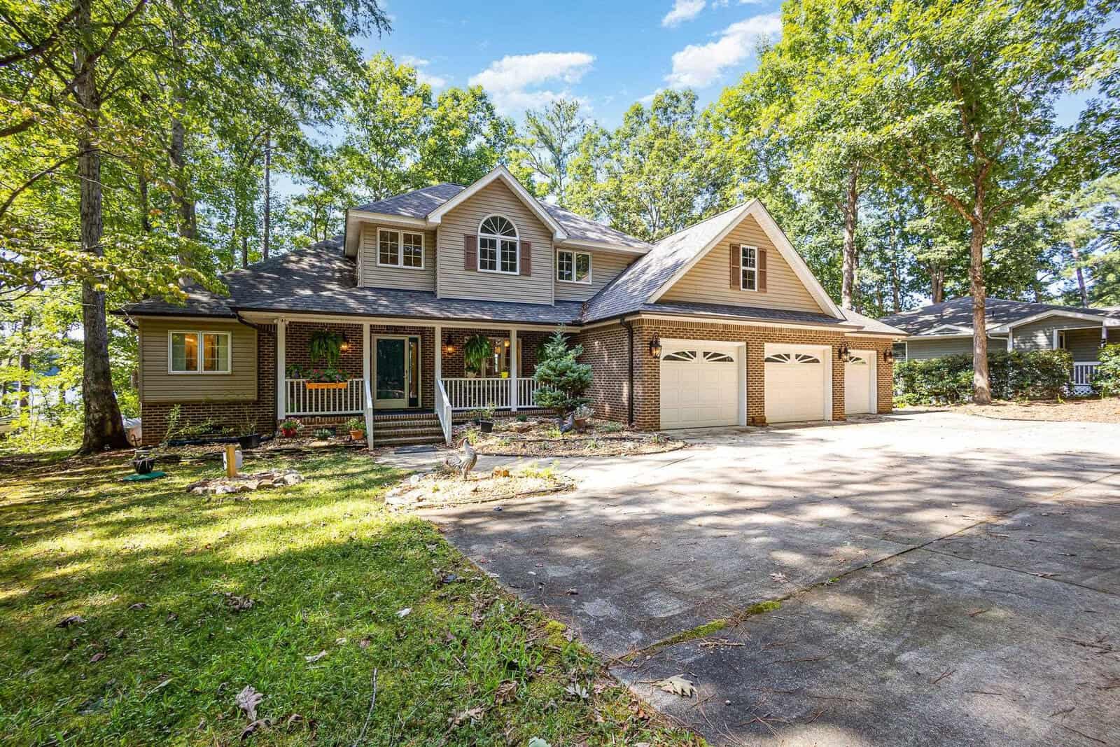 Two-story suburban house with gray siding and brick accents, a large three-car garage, front porch with white railings, and surrounded by tall trees and greenery under a clear blue sky.