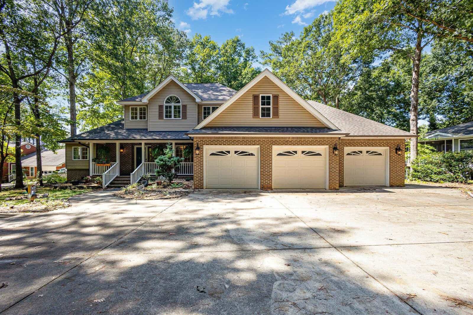 A two-story house with a large driveway, three-car garage, brick and siding exterior, covered front porch, and surrounded by tall trees and greenery under a blue sky.