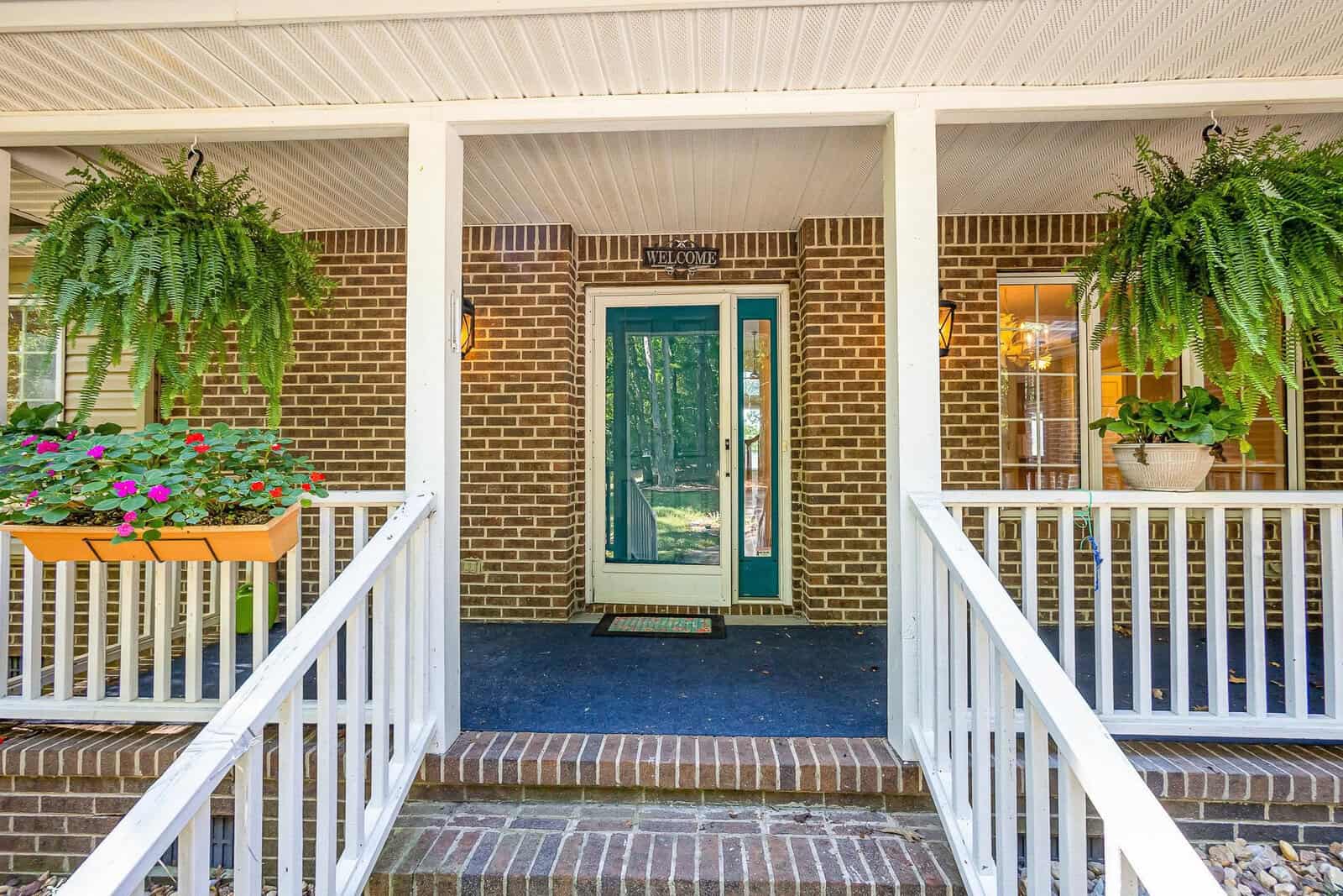 Front porch of a brick house with white railings, a blue-green front door, hanging ferns, a planter with pink flowers, and a potted plant near the door. Steps lead up to the entrance under a covered roof.