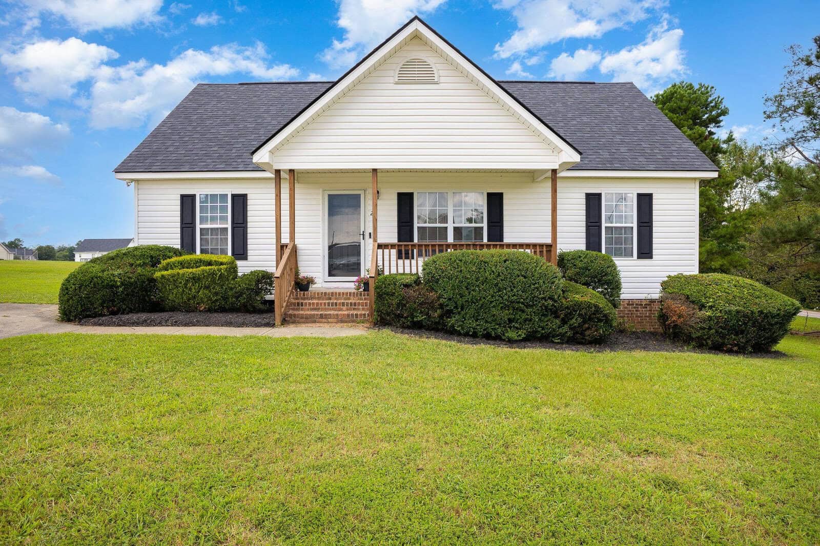 A single-story suburban house with a gabled roof, white siding, and a small front porch, set against a grassy lawn with shrubbery near the foundation.