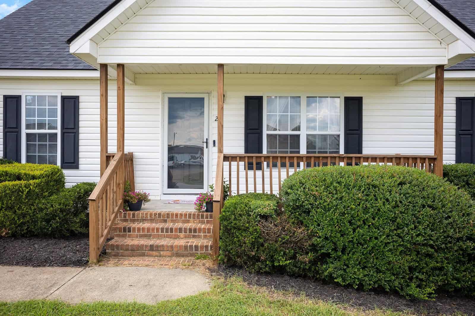 A suburban single-story house with a small front porch, white siding, two windows, and a wooden stairway leading to the front door.