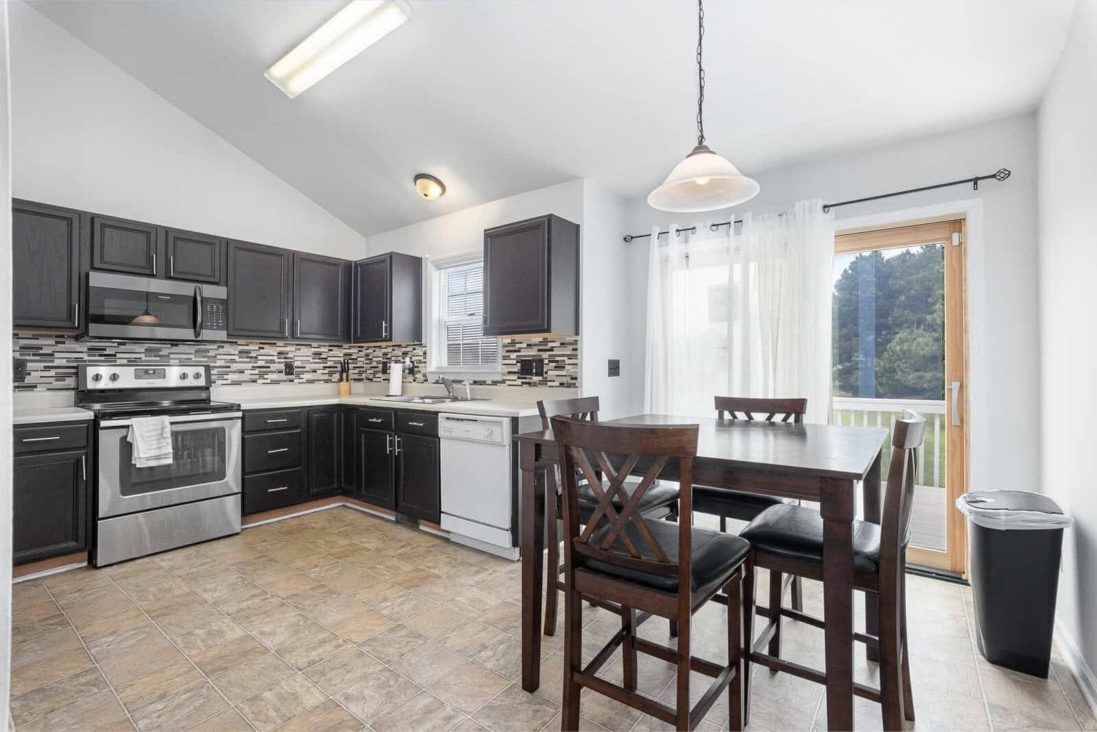 A modern kitchen featuring black appliances, wooden cabinets with dark finish, a tiled backsplash, a center island with bar stools, and a dining area next to a window with curtains.