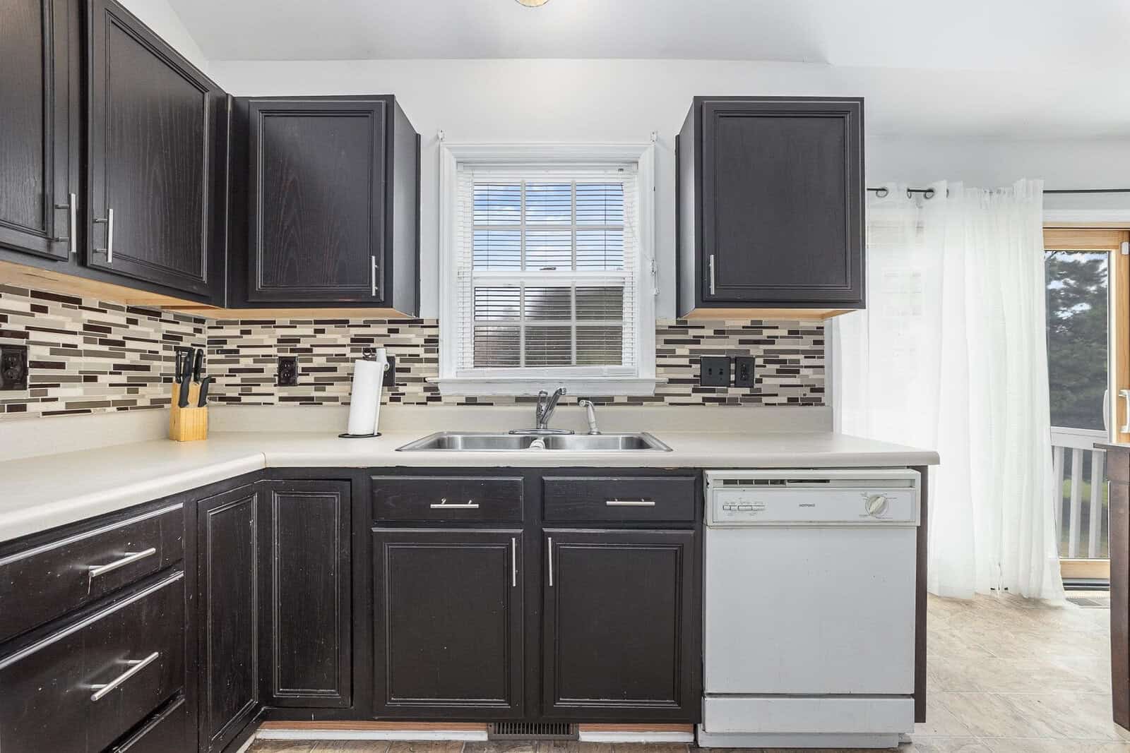 A contemporary kitchen featuring black cabinets, a multi-colored tile backsplash, light countertops, a double sink, a dishwasher, and a window with horizontal blinds, with a doorway leading to an adjacent room that has a white curtain.