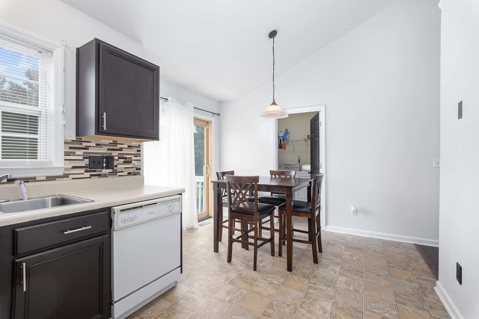 A clean and bright kitchen with white appliances, dark cabinets, a patterned backsplash, and a dining area with a wooden table and chairs under a pendant light.