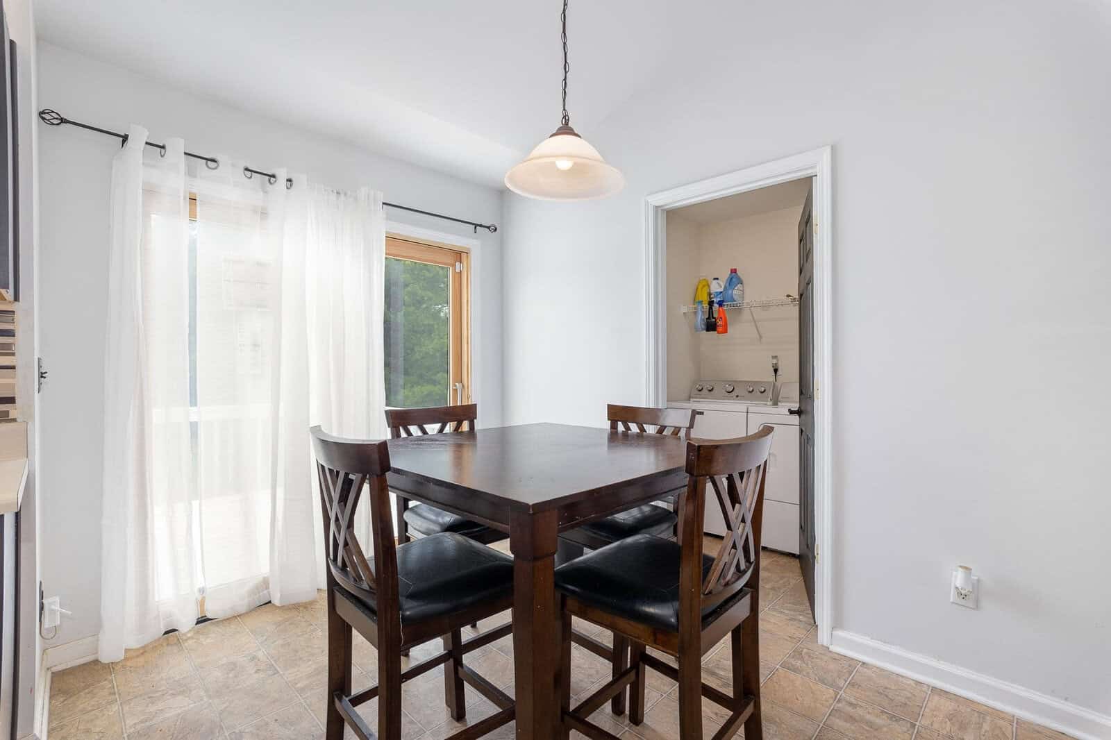 A well-lit dining area with a wooden table, four chairs, white curtains, and a doorway leading to an adjoining room with storage shelves.