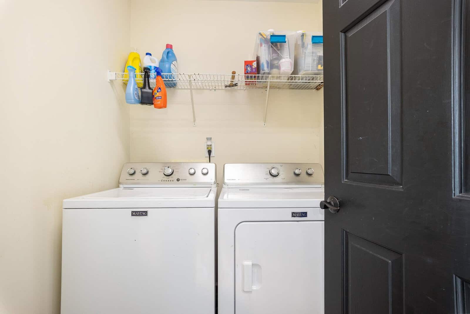 A laundry room featuring a white top-loading washing machine and a dryer, with a shelf above holding various cleaning supplies.