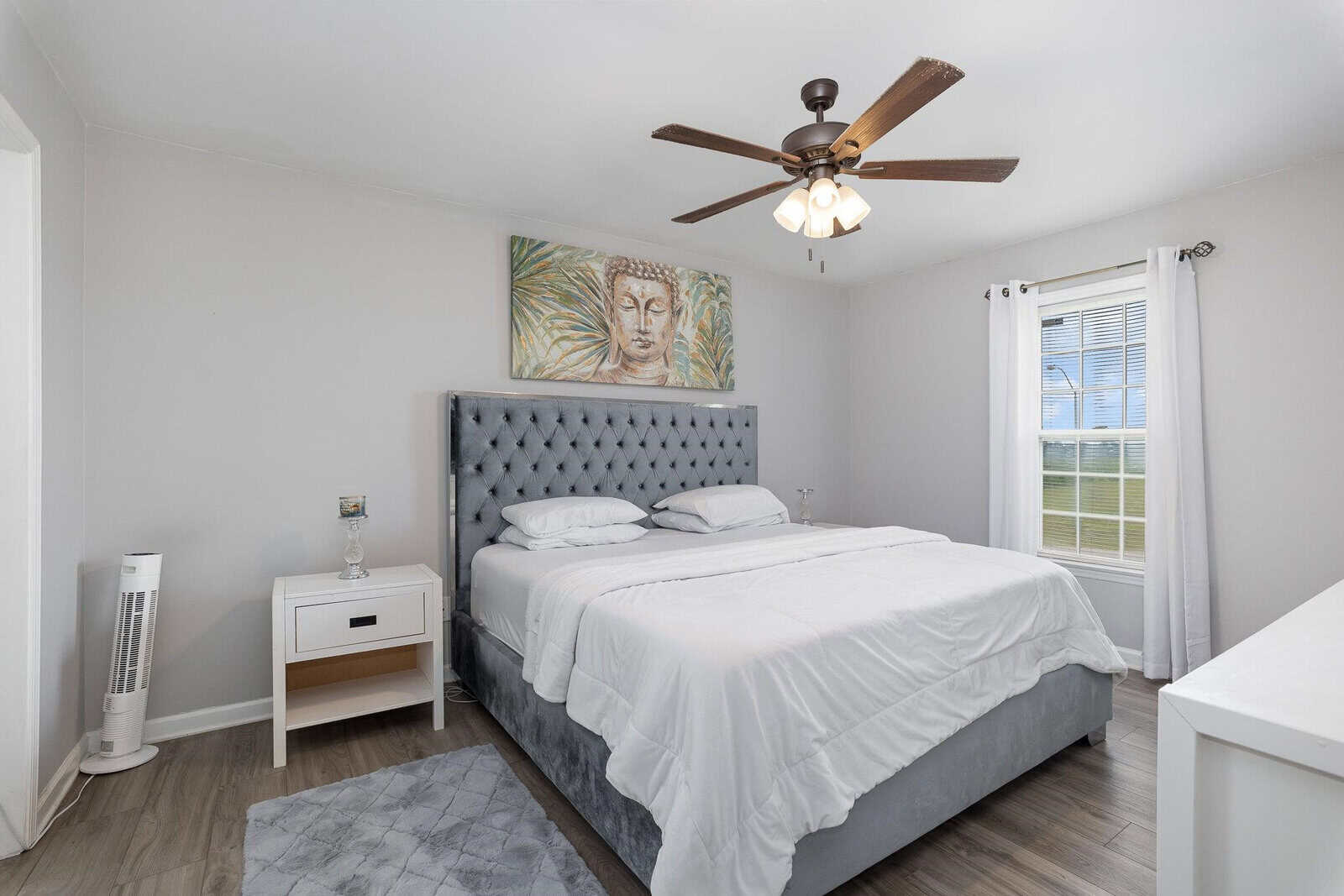 A neatly arranged bedroom with a large bed, gray headboard, white bedding, a ceiling fan above, and a painting hung on the wall.