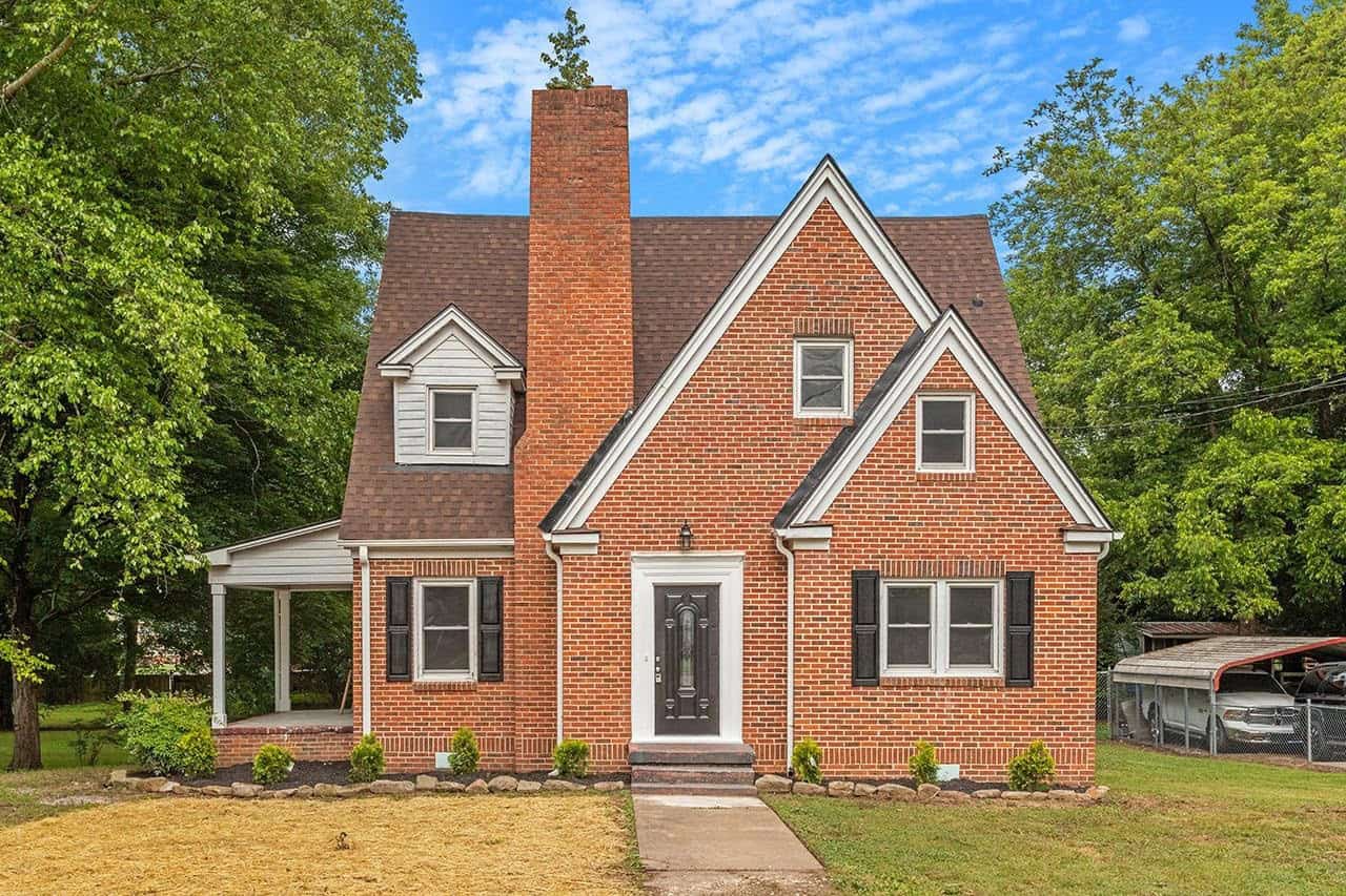 A two-story brick house with a gabled roof and white trim windows, featuring a small front lawn with newly planted shrubs and a concrete walkway leading to the front door.