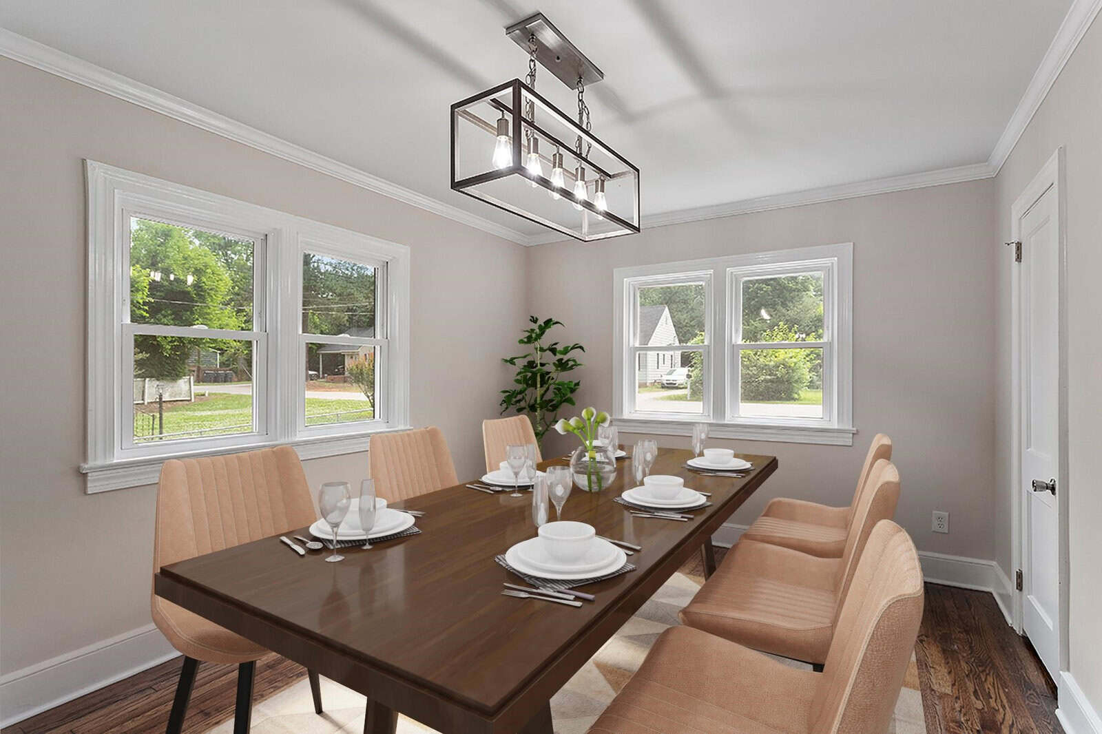 A neatly arranged dining room with a wooden table set for four, beige upholstered chairs, and a contemporary light fixture, surrounded by windows providing ample natural light.