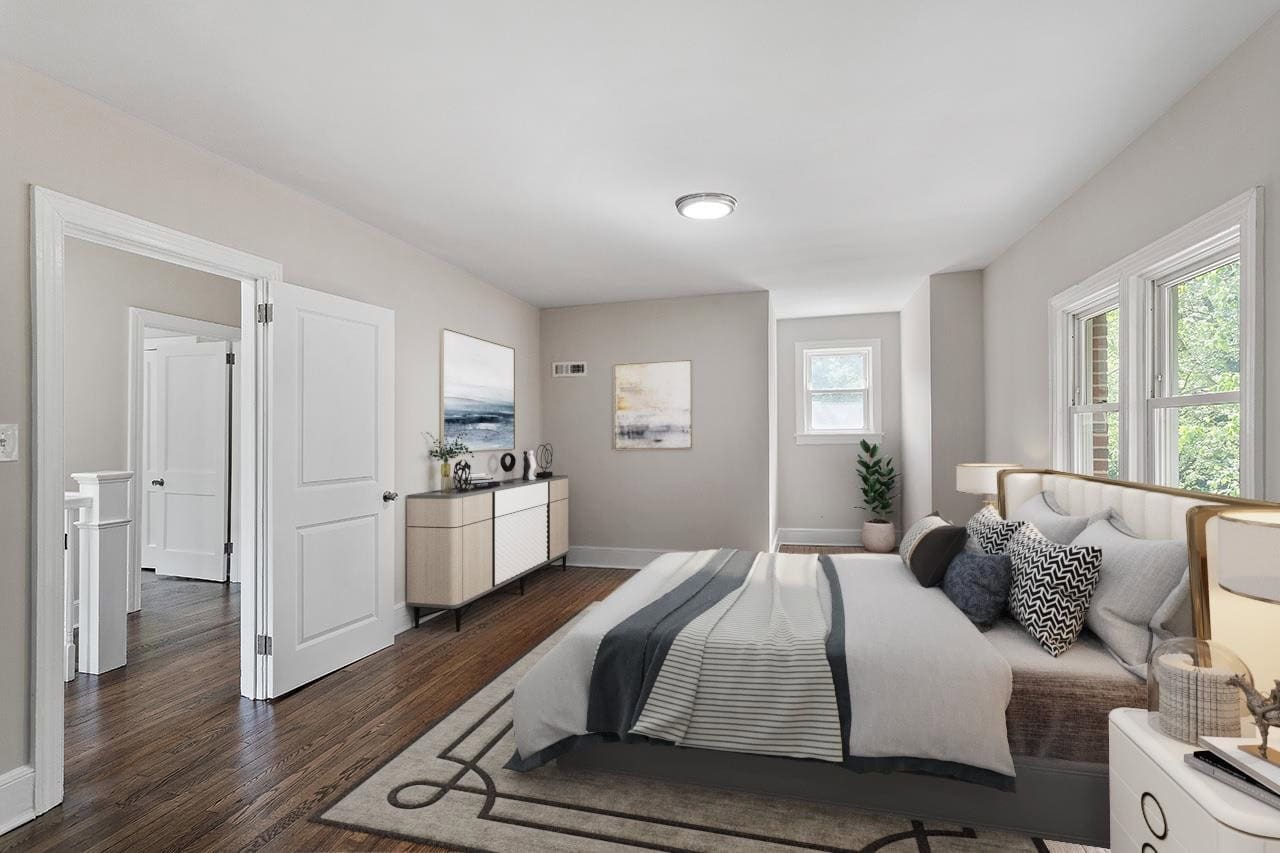 A well-lit, modern bedroom featuring a neatly made bed, hardwood floors, a sideboard with decor, and two windows allowing natural light.