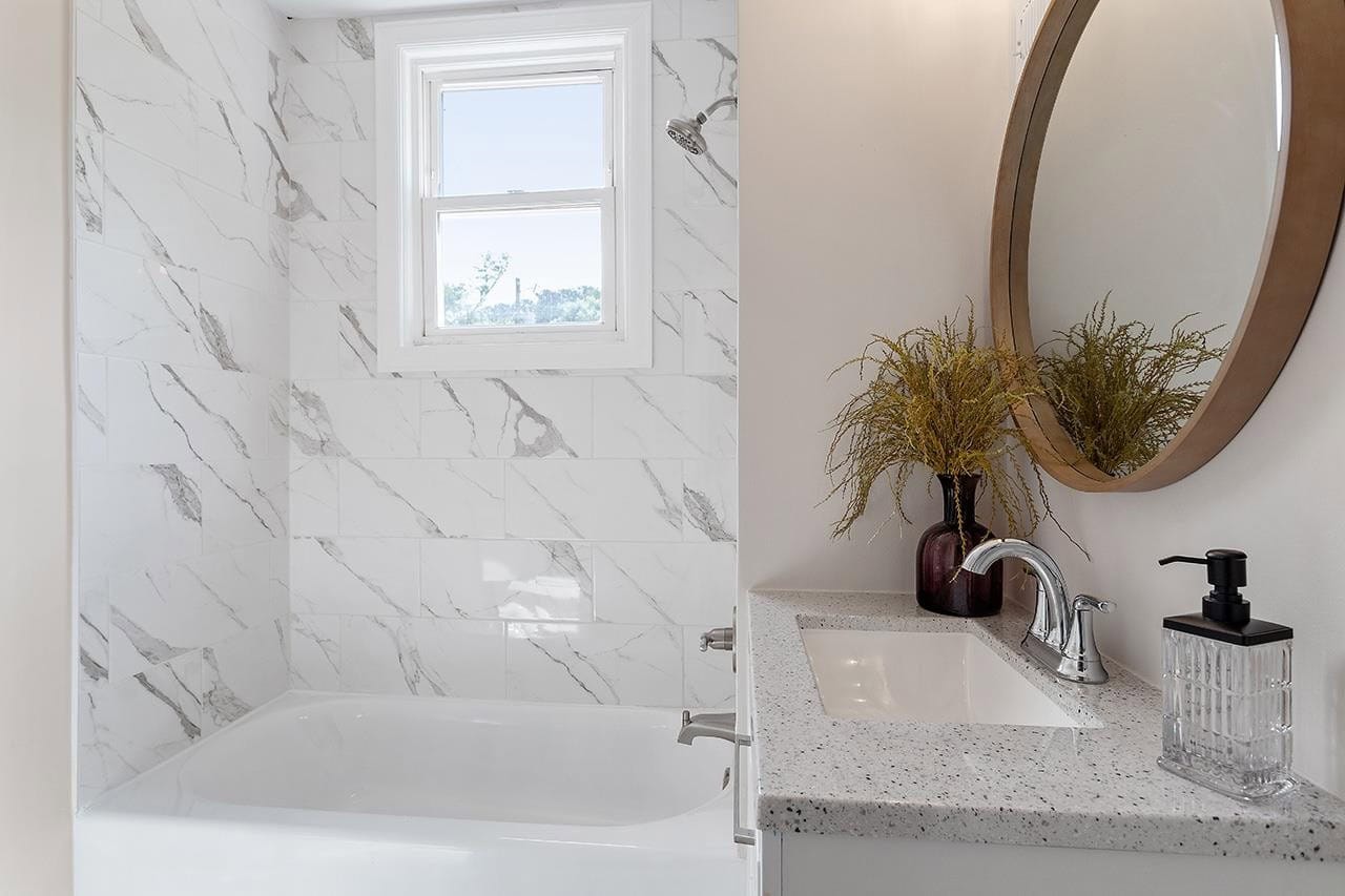 A modern bathroom featuring white marble tile walls, a rectangular bathtub, a granite countertop with an integrated sink, and a round mirror, complemented by a small window and decorative greenery.