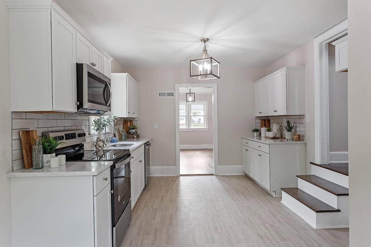 A modern kitchen interior with white cabinetry, stainless steel appliances, and pendant lighting leading to a room with staircase.