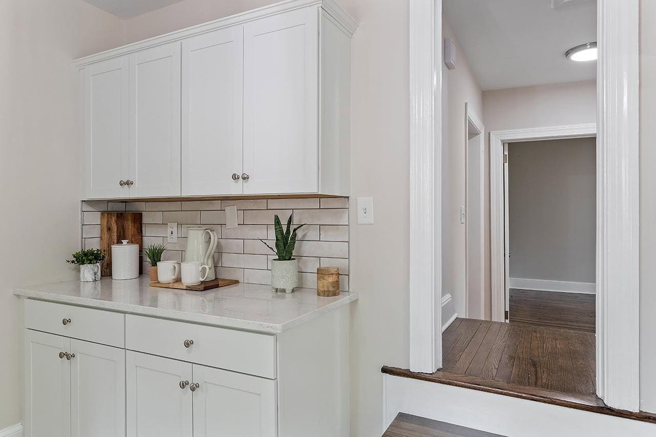 A neatly kept kitchenette with white cabinetry, beige tiled backsplash, and a countertop holding a few decorative items and kitchen utensils, adjacent to a hallway with hardwood flooring.