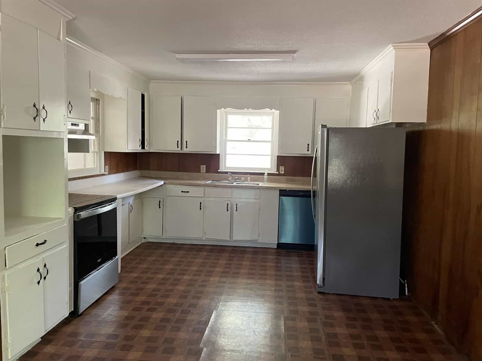 A modest kitchen interior with white cabinetry, a brown floor, and a full-sized refrigerator.