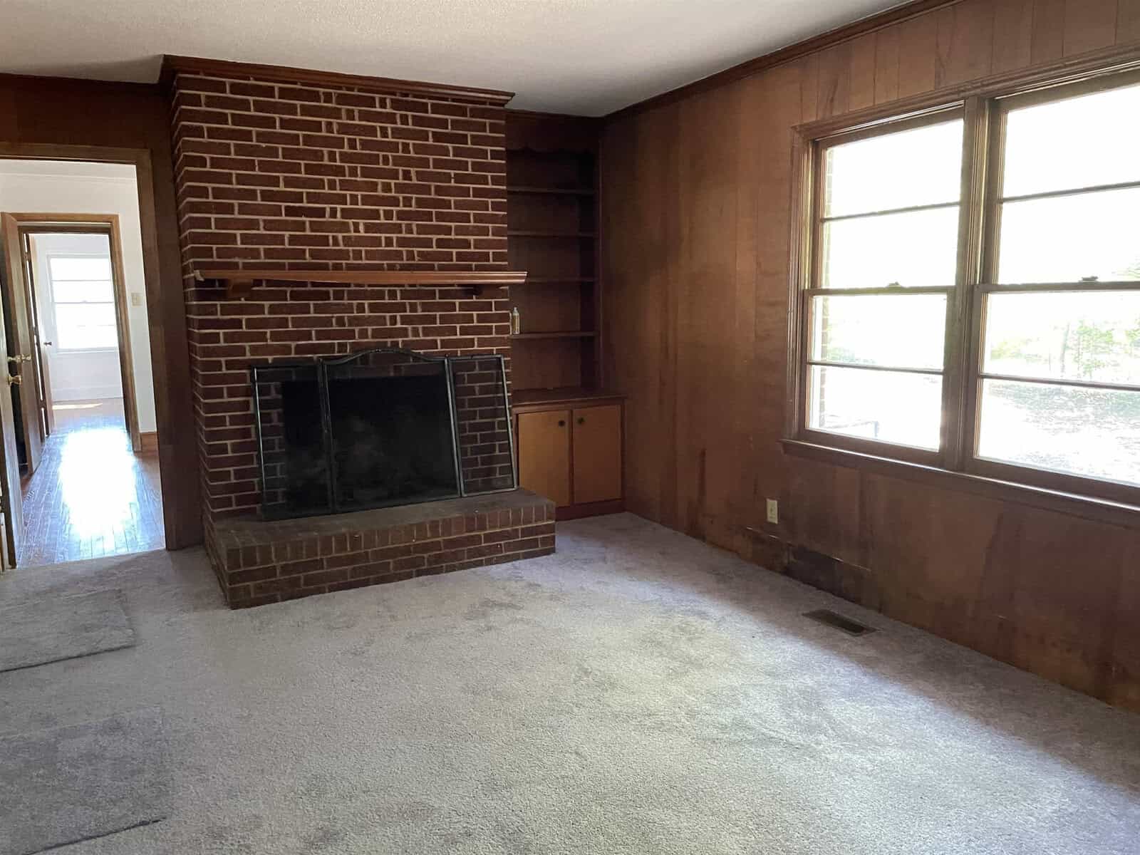 An empty room with a brick fireplace, built-in shelving, carpeted floor, and a large window letting in natural light.