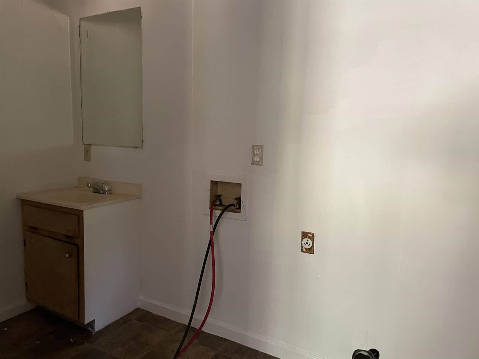 A bare room with white walls featuring a small, simple wooden sink cabinet on the left and electrical outlets on the right, one of which has a red and black cable plugged into it. the room appears to be in a state of disrepair or under renovation.