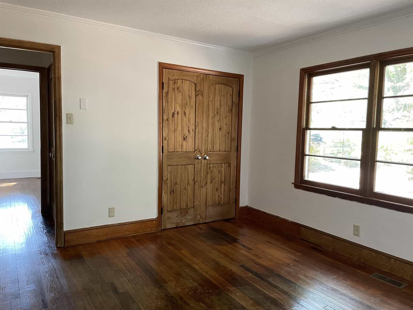 An empty room with wooden flooring, a wooden door, and a window allowing natural light to enter.
