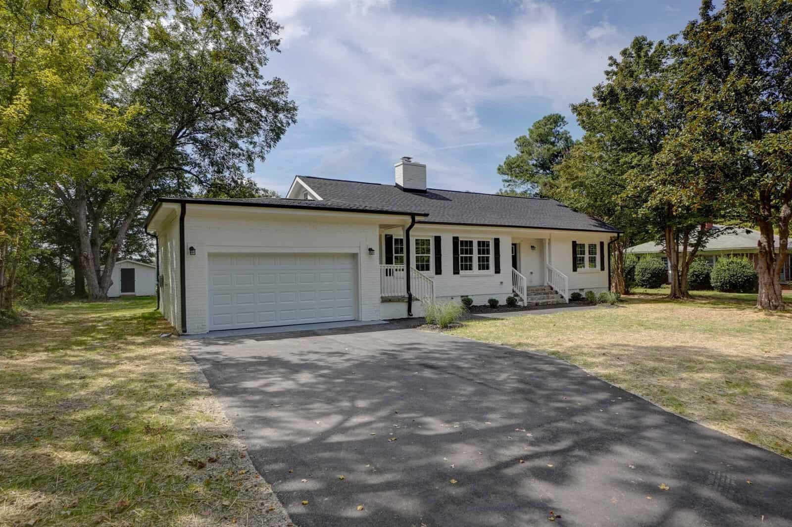 A single-story white house with a garage and an asphalt driveway, surrounded by trees and lawn, under a clear sky.