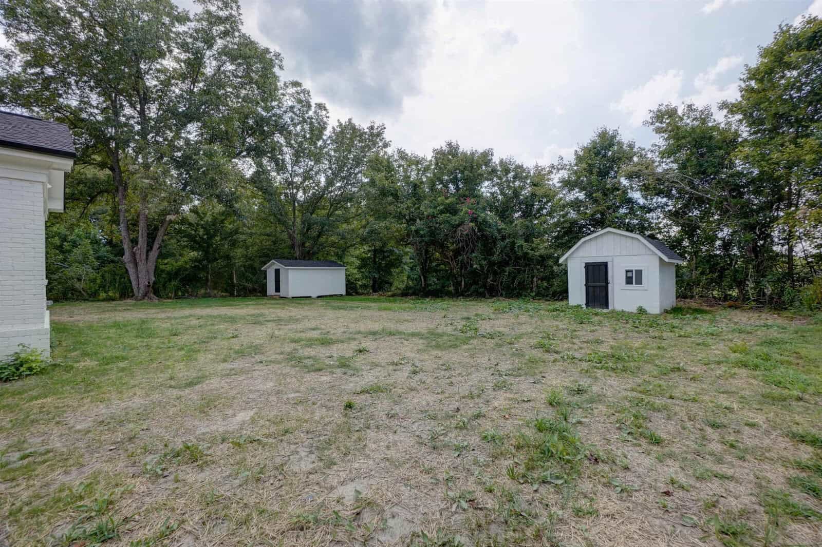 A spacious backyard with two storage sheds surrounded by trees under a cloudy sky.