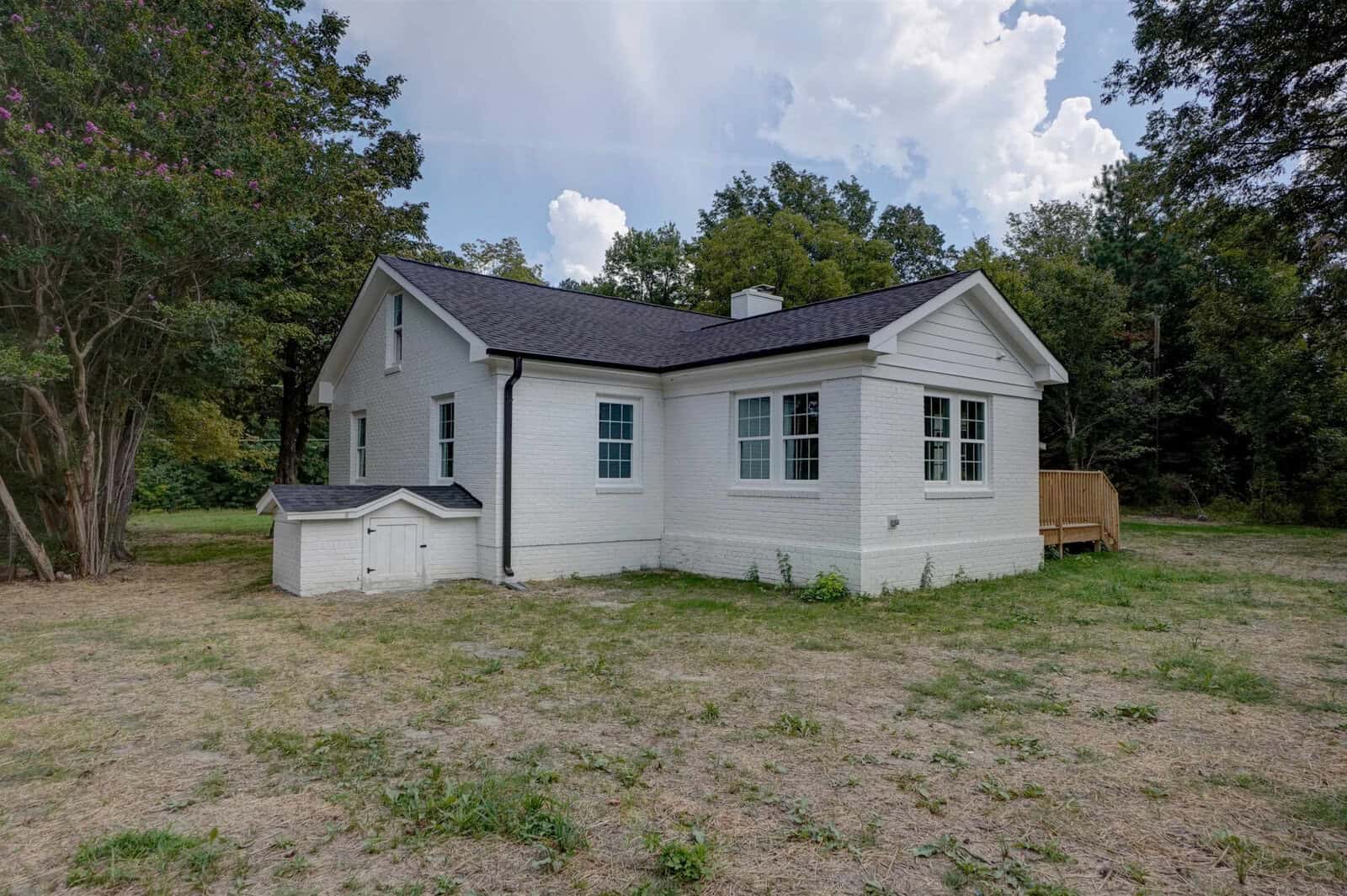 A single-story white house with a small matching doghouse on a grassy lawn with surrounding trees.