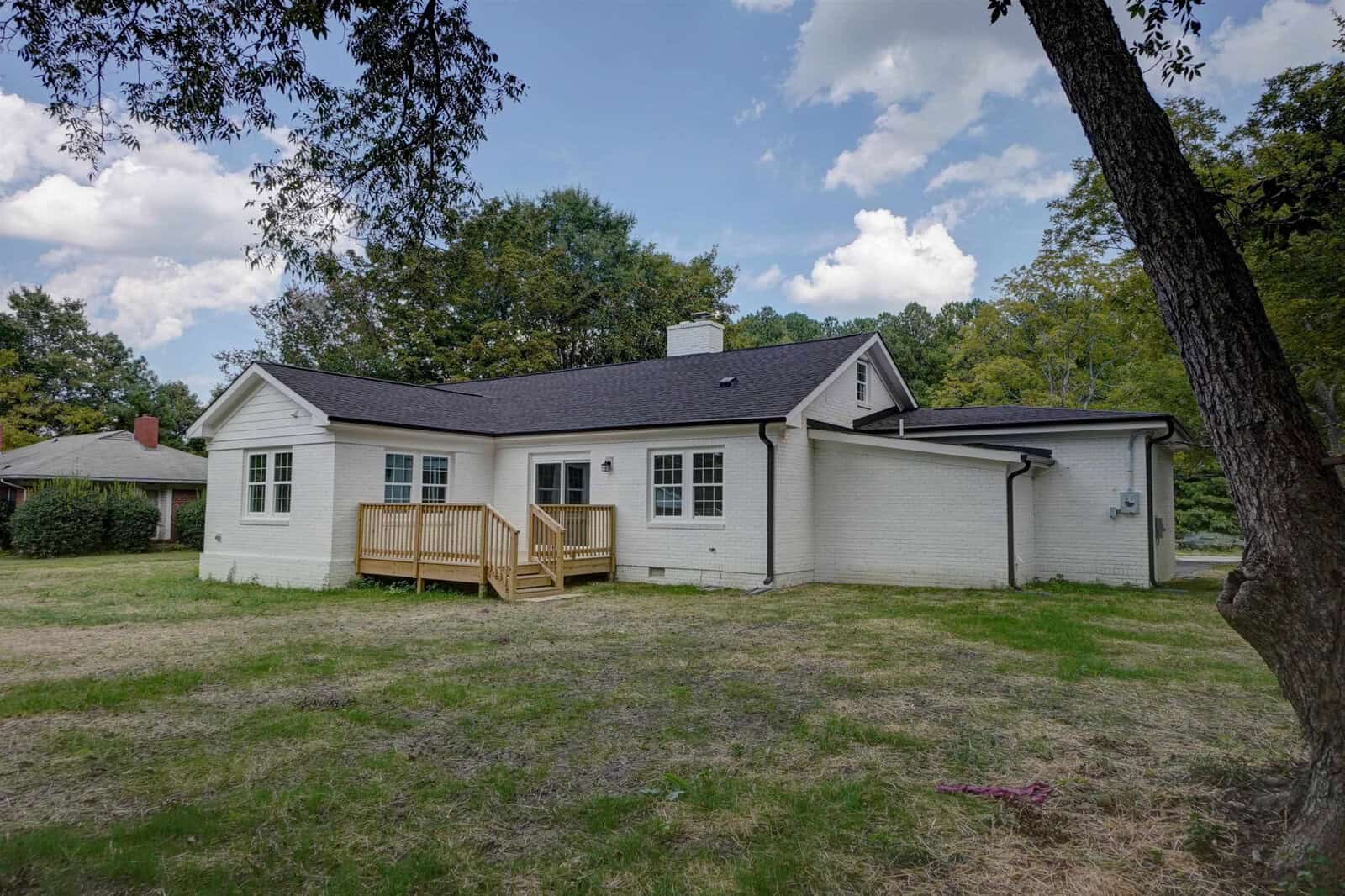 A single-story white house with a gray roof and wooden deck, situated in a grassy yard with a tree on the left.
