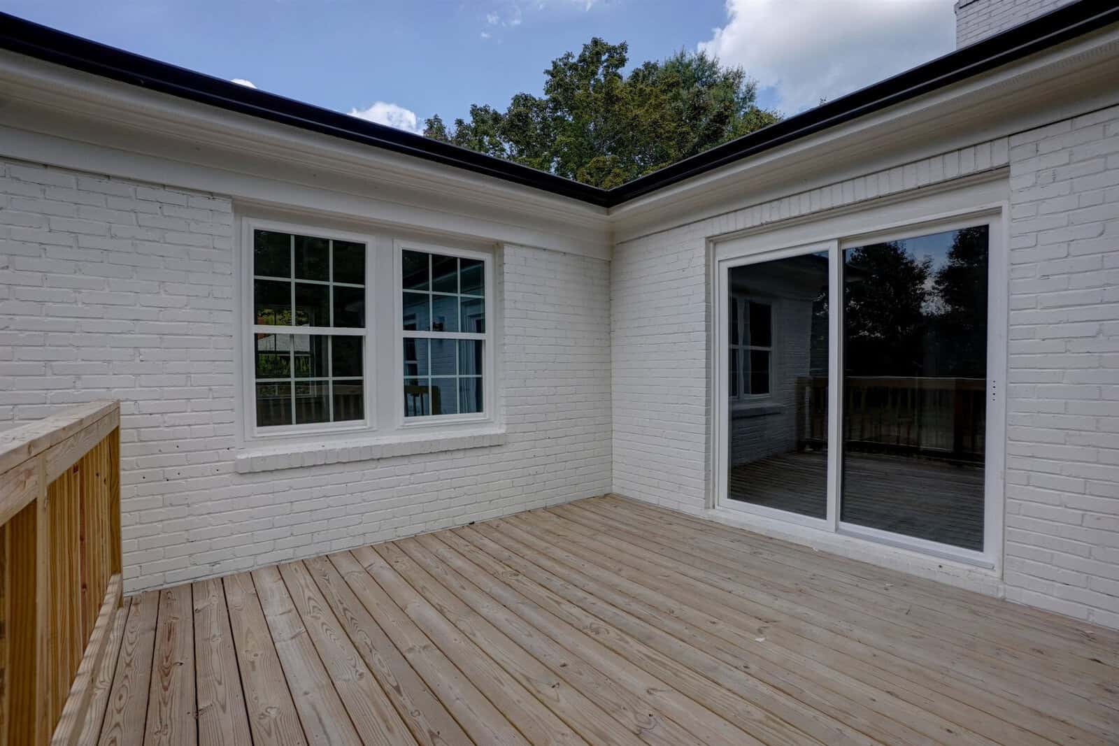 A wooden deck adjacent to a white brick house with multiple windows and a sliding glass door.