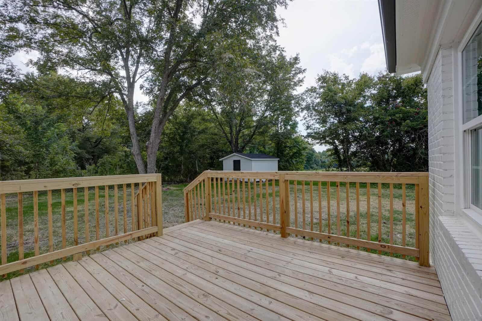 A spacious wooden deck extends from a house with white siding, overlooking a yard with trees and a small shed in the background.