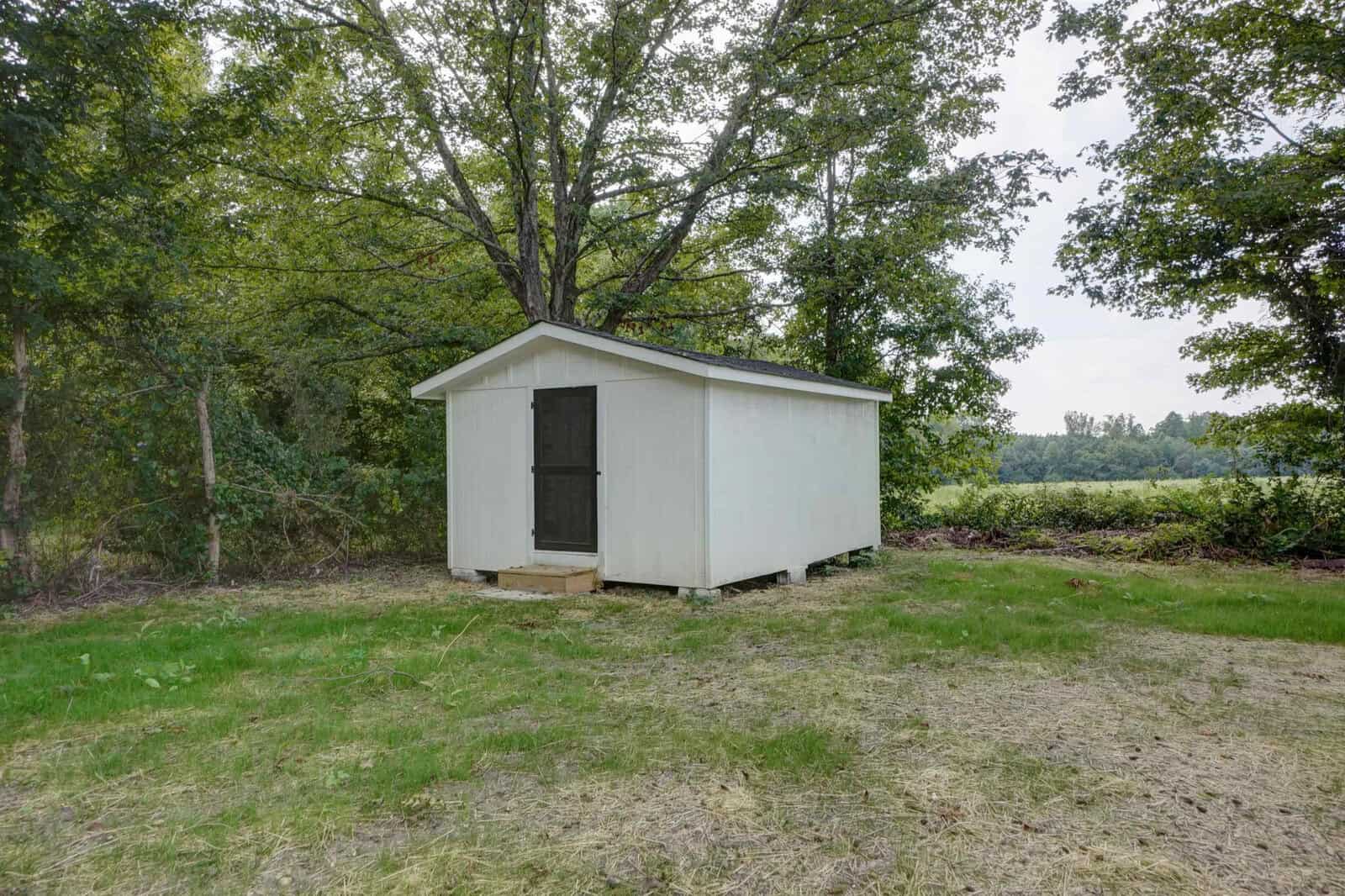 A small, simple white shed with a black door situated in an open grassy area surrounded by trees.