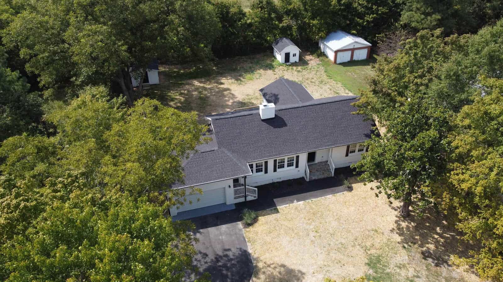 Aerial view of a single-story house with a gray roof surrounded by green trees, with two sheds located in the backyard.