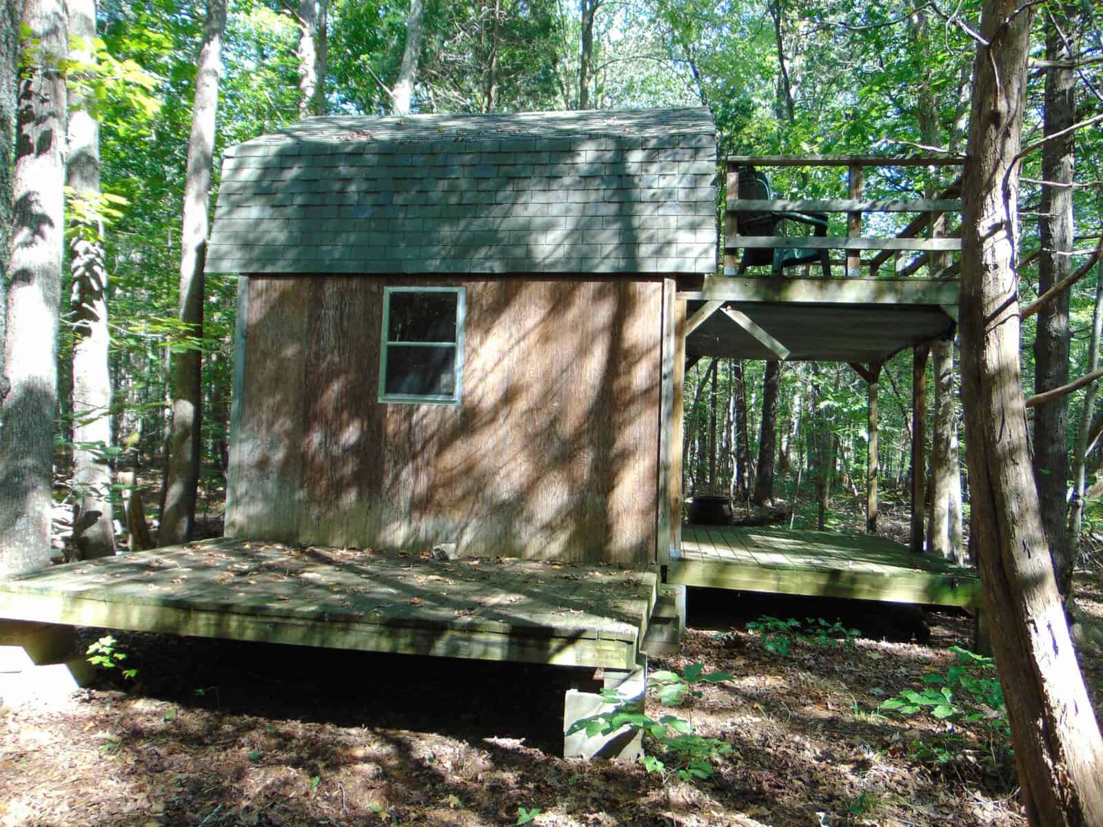A small wooden cabin with a shingled roof and a single window, surrounded by trees, stands on a raised deck in a sunlit forest. Shadows from the trees dapple the cabin and ground.