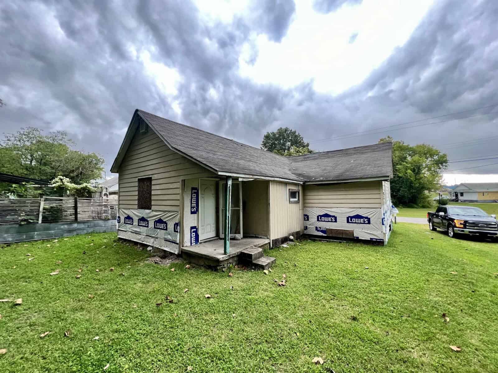 A small, single-story house with beige siding under a cloudy sky, featuring building materials from lowe's on the porch, indicating pending construction or repairs. a vehicle is parked to the right, and the house is surrounded by a lawn.