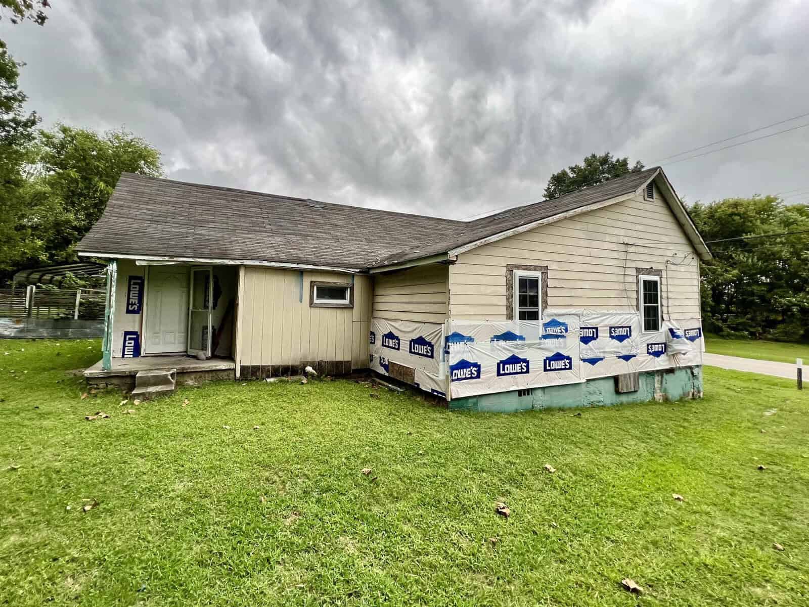 A small, single-story house undergoing renovations, partially wrapped in protective lowe's house wrap, set against an overcast sky.