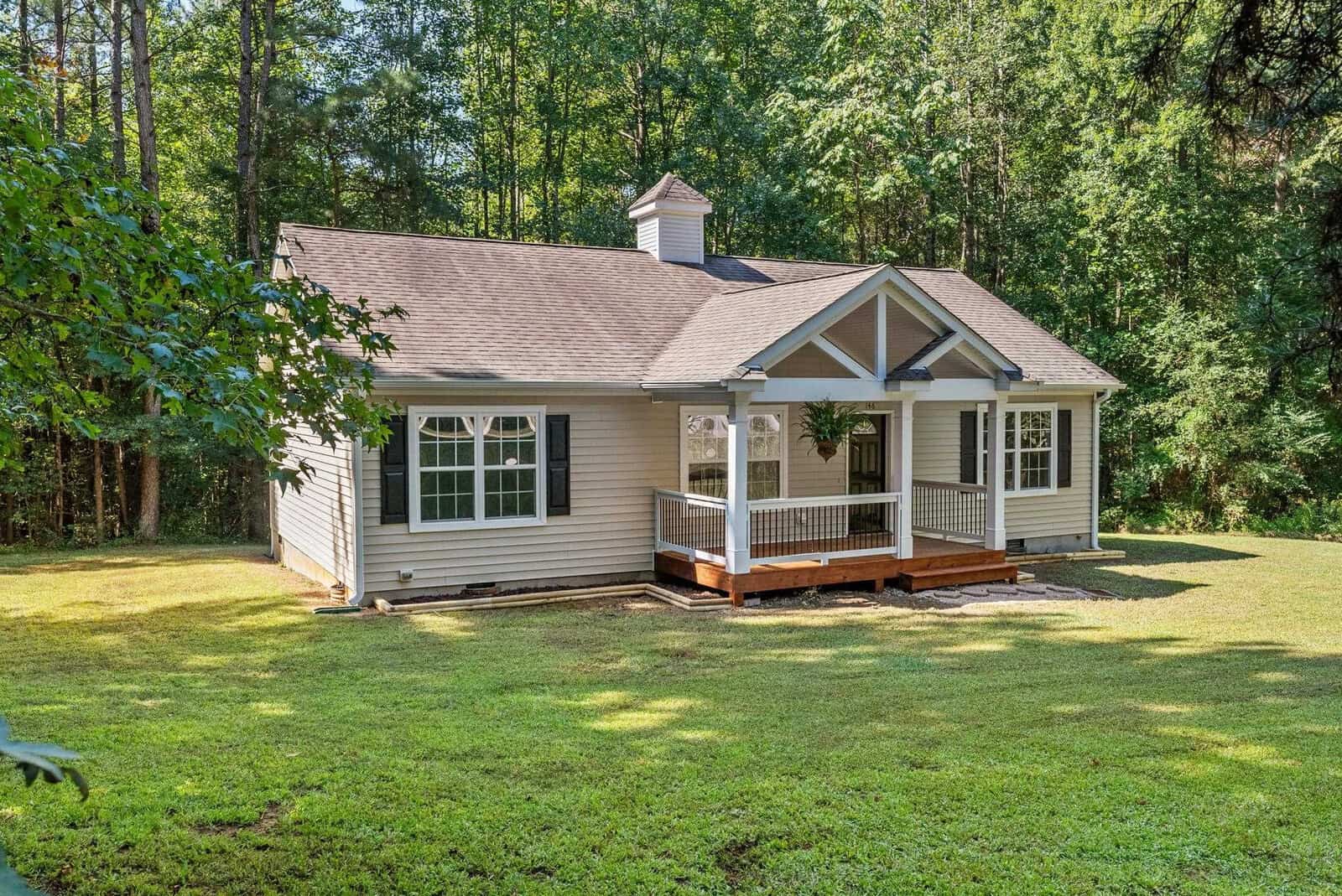 A single-story beige house with white trim and a small front porch sits surrounded by a lush green lawn and trees in a serene, forested setting.