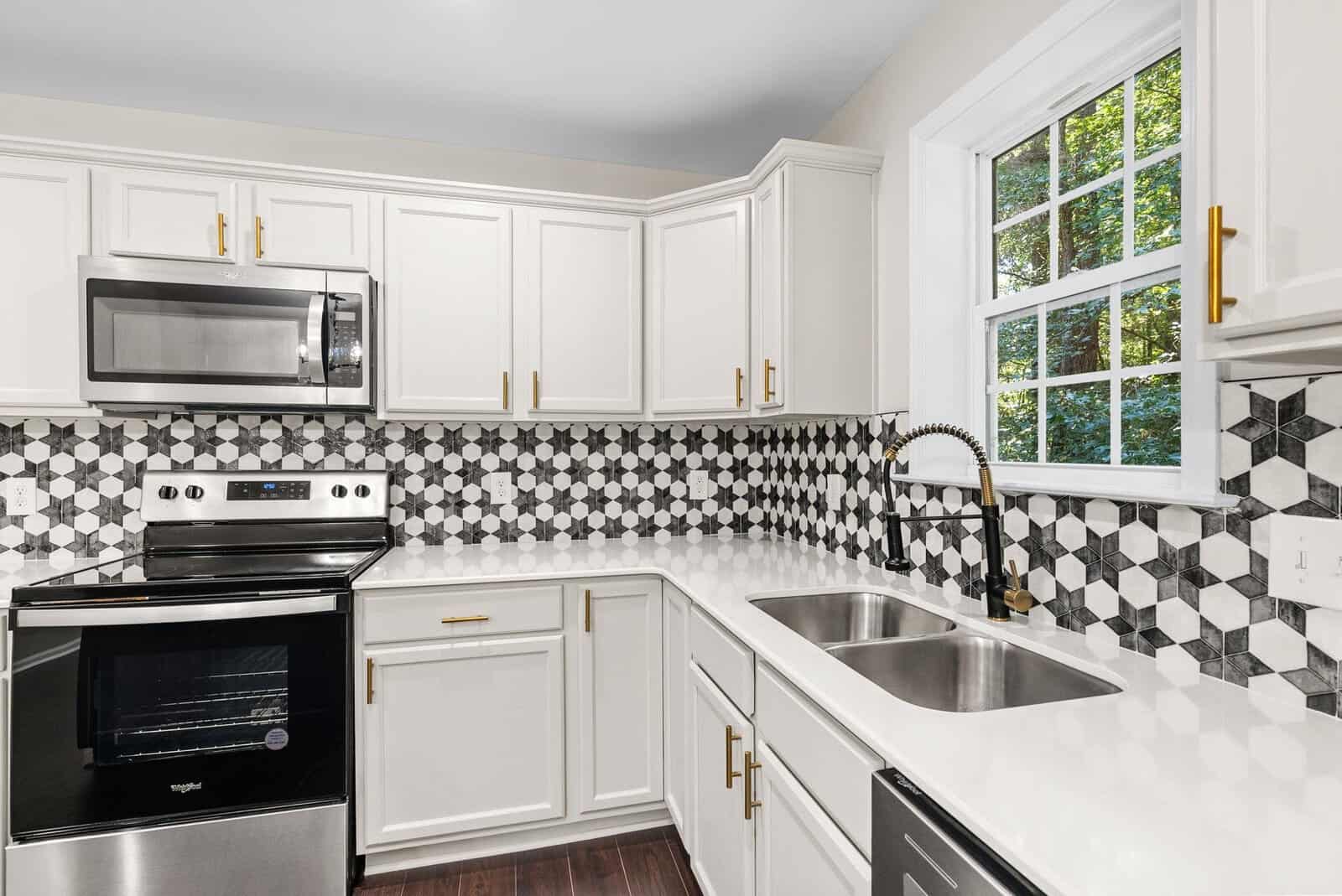 A modern kitchen featuring white cabinetry, black and white backsplash tiles, stainless steel appliances, and gold hardware accents, with a view to the outdoors through a window.