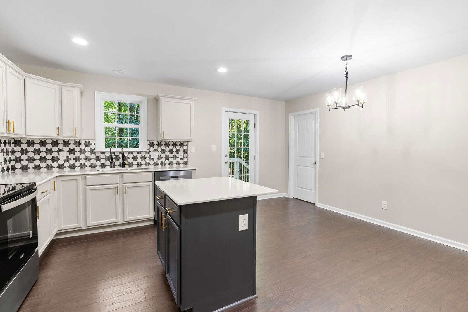 A modern kitchen with white cabinetry, black countertops, and dark wooden flooring, featuring a central island and a view of greenery outside the window.