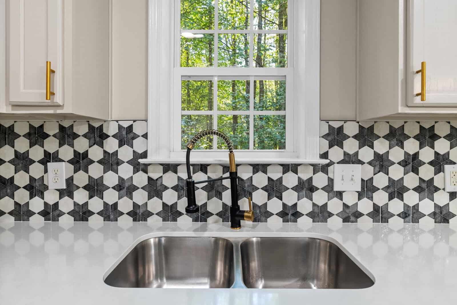 A modern kitchen sink with a black faucet set against white cabinetry, geometric backsplash tiles, and a view of greenery through the window.