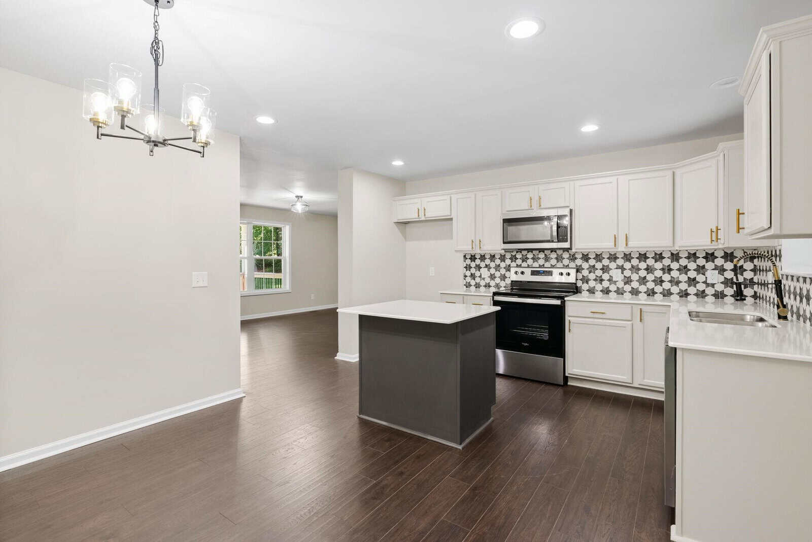 A spacious modern kitchen featuring white cabinetry, stainless steel appliances, a central island, and dark hardwood flooring, illuminated by natural light from the window and elegant overhead lighting.
